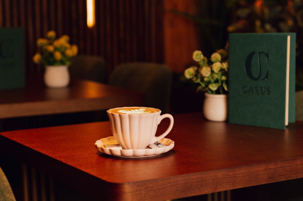 Šolja kafe sa latte art-om, meni Casus i cveće na drvenom stolu.