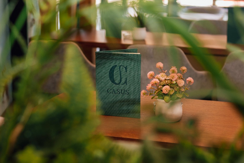 Meni Casus i cveće na drvenom stolu, uokvireno zelenilom.